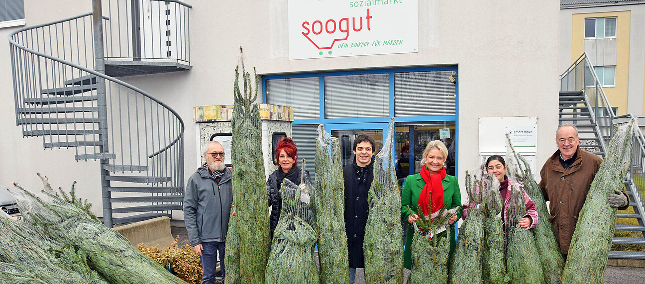 Eine Gruppe von Personen mit Christbäumen vor einem Sozialmarkt.