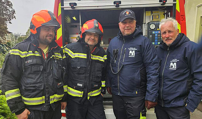 Vier Männer in Feuerwehruniform vor einem Einsatzfahrzeug.